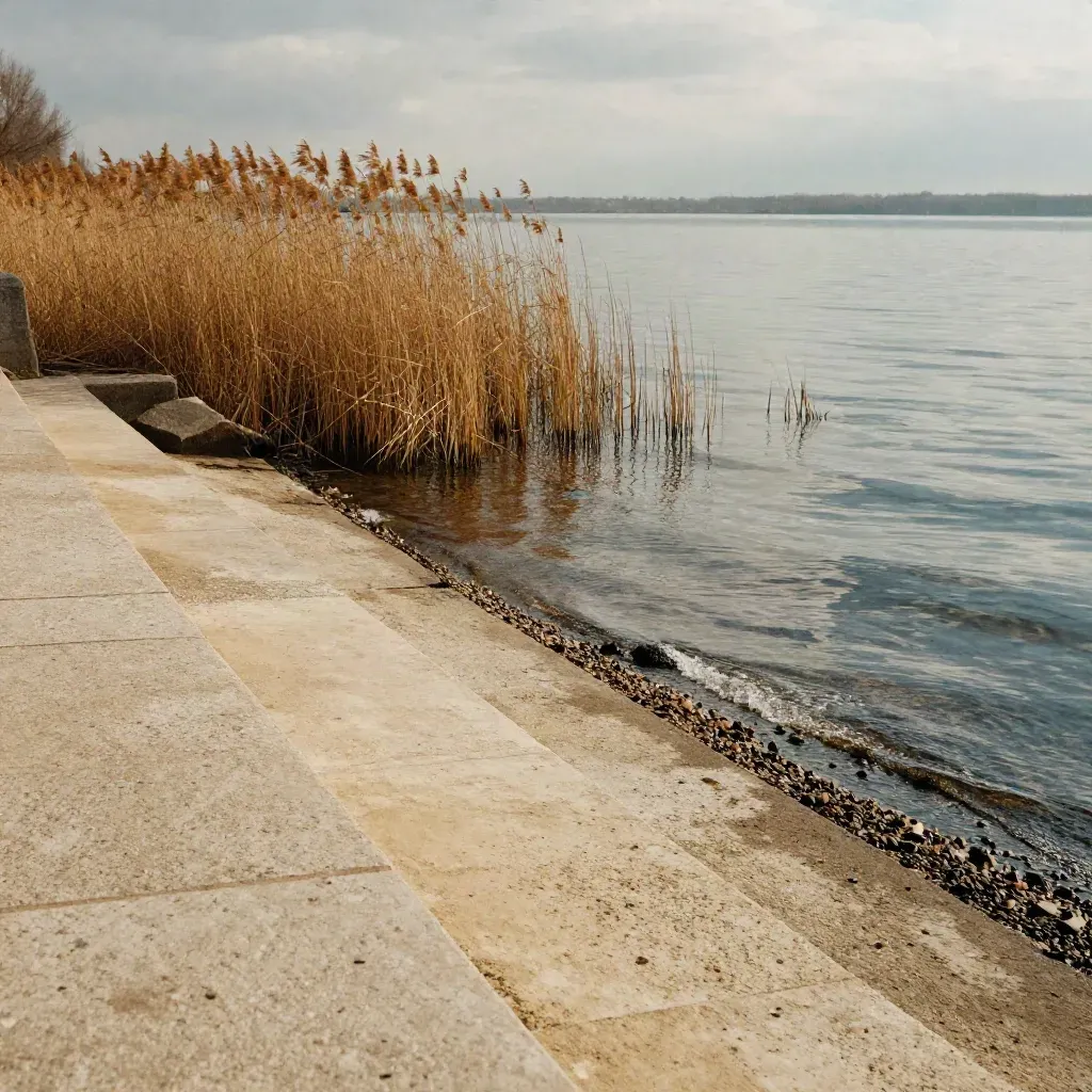 Rive du lac d’Enghien avec muret de pierre claire, roseaux et eau lisse sous ciel voilé.